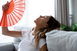 Close up overheated woman waving paper fan, breathing air, leaning back on couch alone, exhausted sick young female feeling unwell, suffering from heating, fever or hot summer weather at home