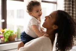 Close up smiling African American woman hugging adorable little daughter, standing at home, beautiful young mum holding cute toddler girl, enjoying tender moment together, childcare concept