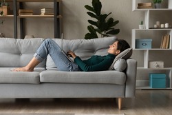 Young Caucasian woman lying on couch in living room work online on laptop gadget. Millennial female relax rest on sofa at home look at computer screen text message on device. Communication concept.