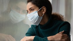 Upset young Caucasian woman in medical facial mask look in window feel lonely distressed sick with covid-19. Unhappy female in facemask suffer from coronavirus. Quarantine, corona pandemic concept.