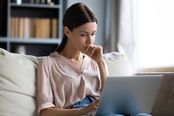 Close up thoughtful upset woman looking at laptop screen, pondering ideas or difficult tasks, sitting on couch at home, pensive young female touching chin, reading news, waiting for message