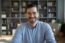 Head shot portrait of happy young handsome confident man in eyeglasses, looking at camera sitting at table at home office. Smiling pleasant businessman holding video call conversation at workplace.