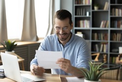 Smiling young businessman in eyeglasses looking at paper correspondence, reading pleasant good news. Excited happy male entrepreneur getting bank loan approvement notification at home office.