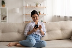 Happy indian lady relax at home alone sit on large couch in comfortable pose share good news at social media via cellphone. Smiling mixed race woman enjoy weekend order goods food online in phone app