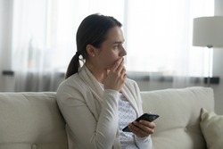 Pensive stressed young Caucasian woman sit on sofa at home using cellphone look in distance pondering of problem. Anxious millennial female feel distressed frustrated with message or text