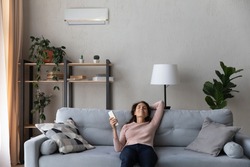 Happy young caucasian woman turning on air conditioner with controller, breathing fresh cooled air or enjoying comfortable temperature indoors, relaxing on comfortable sofa in modern living room.
