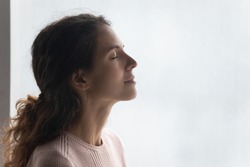 Side head shot view mindful smiling beautiful young woman breathing fresh air, standing near window. Happy millennial caucasian lady enjoying meditation moment, feeling peaceful indoors, copy space.