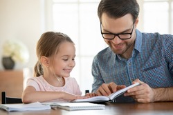 Happy caring young dad and little daughter sit at table at home learn read book prepare for school together. Smiling loving Caucasian father and small girl child study do homework. Education concept.