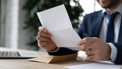 Crop close up businessman sit at desk reading post paper letter or correspondence in office. Male employee or director receive consider postal paperwork or mail notification at workplace.