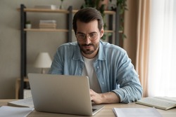 Attentive millennial male manager employee busy at telework at quarantine time study documents in corporate network from home pc. Concentrated young man specialist doing remote computer work on laptop