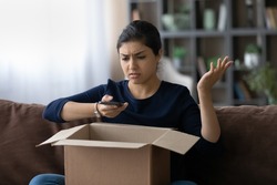 Unhappy young Indian woman frustrated by wrong order shopping online using smartphone. Upset ethnic female buyer unbox parcel buying on web confused with bad delivery service. Shipping concept.