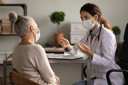 Close up serious doctor wearing medical face mask consulting mature woman patient at appointment in office, physician explaining treatment, giving recommendations, elderly generation healthcare