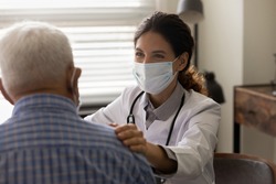 Close up smiling female doctor wearing protective face mask touching senior patient shoulder, physician comforting and supporting mature man at medical appointment, psychological help concept