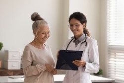 Head shot close up smiling female doctor wearing glasses and white uniform consulting mature patient about treatment, holding clipboard, happy senior woman and physician standing in hospital
