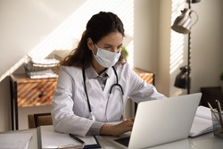 Close up focused professional female doctor physician wearing protective face mask using laptop, looking at screen, consulting patient online, chatting, browsing medical apps, searching information