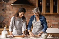 Happy Latino senior mother and grownup daughter make dough prepare sweet pie or pastries at home on weekend. Smiling Hispanic mature mom and adult girl cooking baking in kitchen. Hobby concept.