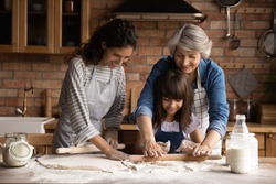 Smiling three generations of Hispanic women have fun baking together with dough at home kitchen. Happy little Latino girl child with young mom and senior grandmother cook pastries or cookies.