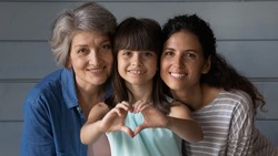 Close up family portrait of happy three generations of Hispanic women pose together show love heart hand gesture. Smiling little Latino girl child with young mother and senior grandmother feel united.
