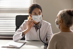 Close up serious female doctor wearing face mask and coat checking senior patient lungs, listening to heartbeat, mature woman breath, elderly generation healthcare, medical checkup concept