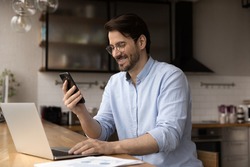 Close up smiling businessman wearing glasses using laptop and phone, looking at smartphone screen, working on online project, financial statistics, chatting with business partner, consulting
