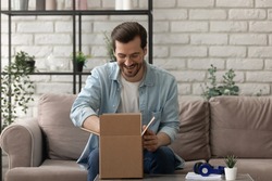 Unpacking parcel. Happy young man postal delivery service client sit on couch at living room open small carton box enjoy goods received. Curious guy online shopper get new order from web store by mail