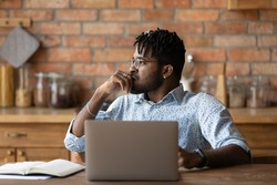 Thoughtful african male freelancer sit by laptop look aside ponder on business problem. Pensive young black man marketing expert work from home analyse sales stats plan creative concept for new brand