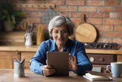Modern day pensioner. Smiling mature latina woman sit at home office by kitchen table watch video on tab computer screen. Old age grandmother enjoy reading e book on tablet pc scroll news on websites