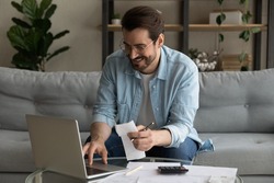 Smiling man wearing glasses using laptop, calculating domestic bills, planning, managing budget, sitting on couch at home, happy businessman browsing online banking service, satisfied by money refund