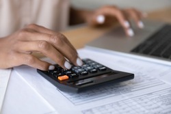 Crop close up of biracial woman calculate expenses expenditures on calculator machine, pay bills taxes online on computer. African American female manage budget on laptop. Saving, finance concept.