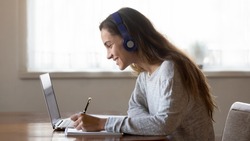 Smiling millennial teen female in headphones look at laptop screen write study online at home. Happy young Latin woman in earphones handwrite make note work distant on computer. Education concept.