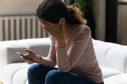 Out of the blue. Frustrated millennial latin woman sit on sofa read sudden bad news from cell screen cover face with palm in desperate gesture. Stressed young lady get nasty blackmail message on email