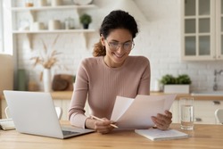 Happy millennial hispanic lady review printed paper documents financial reports satisfied with good state of business low tax rate paid debt confirmation. Glad young female do paperwork read good news