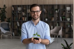 Portrait of smiling young businessman hold soil and small plant launch startup project or activity. Happy millennial male employee or CEO with seedling sprout in hands. Growth, development concept.
