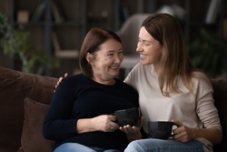 Smiling grownup Caucasian female kid child relax rest on couch with mature 60s mom drink coffee tea together. Happy two generations of women enjoy family reunion at home. Relation concept.