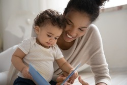 Happy young African American mom play read book to cute little baby child relax together at home. Smiling loving ethnic mother enjoy motherhood with small biracial toddler kid. Parenthood concept.