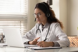 Modern medicine. Focused young female doctor general practitioner in headset watching medical webinar video conference on laptop screen taking notes. Young woman medic intern learning online using pc