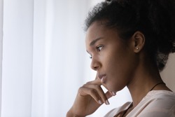 Close up head shot side view young african american thoughtful woman standing near window, looking in distance, feeling puzzled with personal problems, pondering about difficulties, copy space.