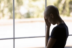 Depressed cancer patient losing hope for remission and recovery, feeling tired, desperate and disappointed. Sad hairless ill young woman standing at window. Oncology treatment concept