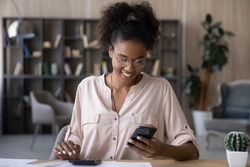 Happy young african ethnic woman in glasses calculating domestic utility bills taxes, managing monthly budget, paying for services online in mobile shopping application, satisfied with enough money.