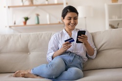 Happy millennial Indian female buyer or client shopping online on smartphone with credit debit card. Smiling young mixed race woman have fun buy pay on internet using secure bank service on cellphone.