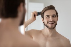 Happy handsome young shirtless man combing smooth straight hair, looking in mirror, enjoying beauty care activity, satisfied with haircare cosmetic products, haircut, barber work result
