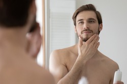 Happy handsome young man applying lotion or balm on stubble after-shaving or trimming, looking at mirror, touching beard. Guy enjoying male beauty care bathroom activity, holding cosmetic flask