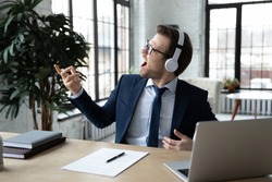 Excited happy funny businessman in wireless headphones listening to music, enjoying favorite song, playing air guitar, pretending to be guitarist, rock star, relaxing and having fun during work break