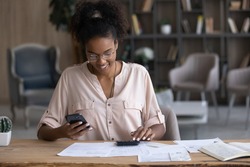 Smiling African American woman in glasses calculating bills, using smartphone and calculator, positive young female browsing online banking service, analyzing financial documents, planning budget