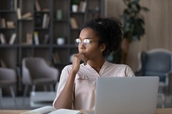 Thoughtful African American woman in glasses looking to aside touching chin, pondering project strategy or ideas, pensive young female student or freelancer sitting at work desk with laptop