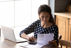 Focused young woman using calculating taxes, budget, costs, paying bill, rent or mortgage fee, checking paper invoice, receipt. Accounting professional with laptop and calculator working from home