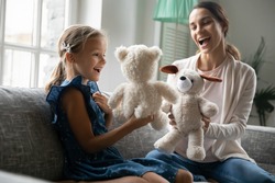 Playful happy young Caucasian mom and small teen daughter have fun play puppet theatre at home. Smiling mother and little girl child engaged in playful game activity with teddy bear toys together.
