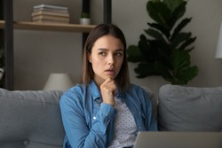 Pensive young woman sit on couch looking deep in thoughts touch chin with hand while thinks over freelance task working remotely from home on laptop. Thoughtful female studying use modern tech concept