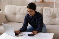 Confident Indian woman calculating bills, paying online, using laptop and calculator, sitting on couch at home, focused young female managing planning household budget, preparing financial report