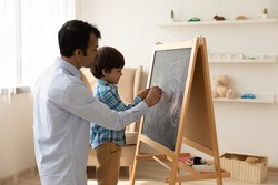 Loving Indian father with little son drawing on black board with colored chalks together, painting, young dad and preschool boy child having fun at home, family involved in creative activity